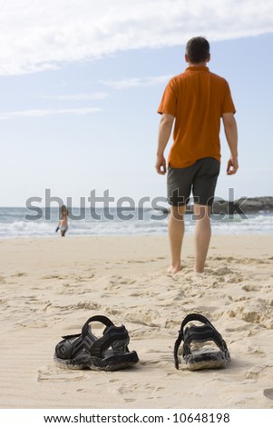 Man walking barefoot on a beach. Woman in the background. Focus on the sandals in the foreground.