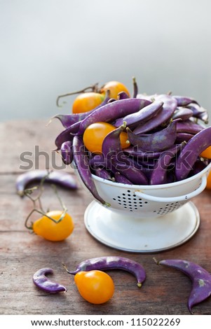 Fresh purple beans and yellow tomatoes in the white colander on wooden surface
