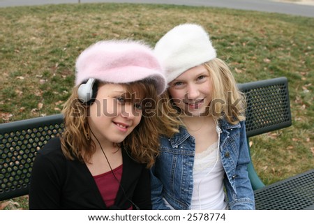 young girl friends smiling and sitting on bench at park listening to music