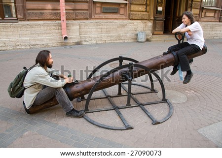 Young smiling couple sit on teeter-totter on street in summer