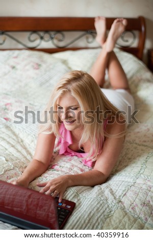 Young sexy blonde woman with computer on the bed smiling and chatting with friend online in her house