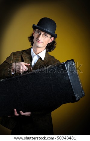 portrait of young english retro gentleman in bowler hat holding suitcase as a luggage. yellow background
