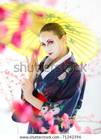 Artistic portrait of japan geisha woman with creative make-up near sakura tree in kimono with umbrella