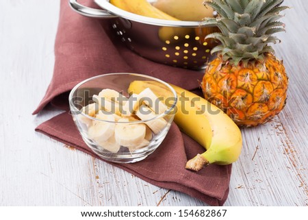 Sliced banana in bowl and pineapple on white wooden background. Selective focus.