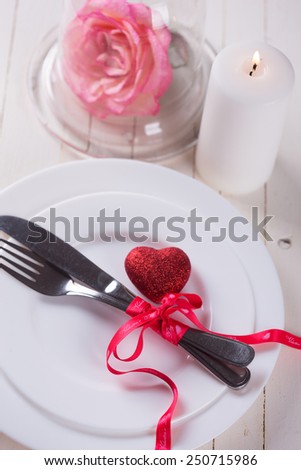 Romantic table setting. White plates, fresh rose in clochet, candle and little red heart.  Selective focus is on bow on ribbon.