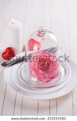 Romantic table setting. White plates, fresh rose in cloche, candle and little red heart.  Selective focus.