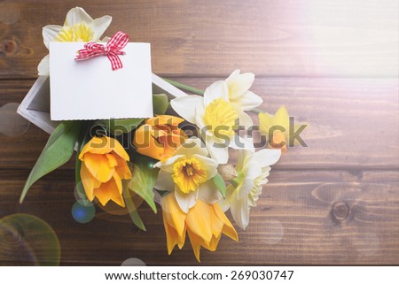 Fresh  spring yellow narcissus and  tulips flowers in wooden box and empty tag in ray of light  on brown  painted wooden planks. Selective focus. Place for text.
