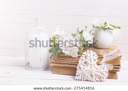 Postcard with apple blossom, decorative heart, old books and candle in decorative bird cage in ray of light on white painted wooden planks. Selective focus.