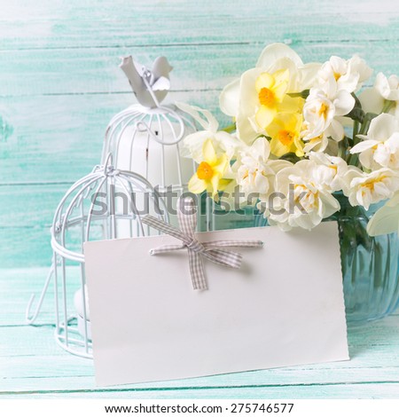 Background with colorful narcissus flowers in blue vase, candles  and empty tag for text on turquoise painted wooden planks. Selective focus. Place for text. Square image.