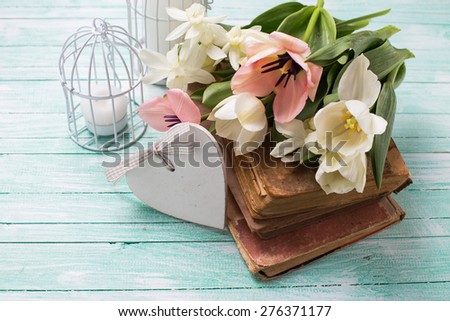 Fresh  spring yellow tulips and narcissus flowers on old books, decorative heart, candles in bird cages on turquoise  painted wooden background. Selective focus.
