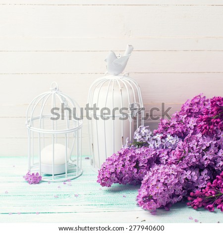 Background  with fresh lilac flowers and candles in decorative bird cages  on turquoise painted wooden planks against white wall. Selective focus. Place for text. Square image.
