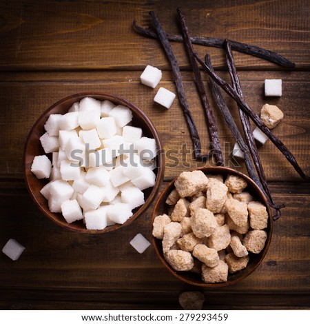 White and brown  sugar cubes in bowls  and vanilla beans on dark painted wooden background. Selective focus. Square image.