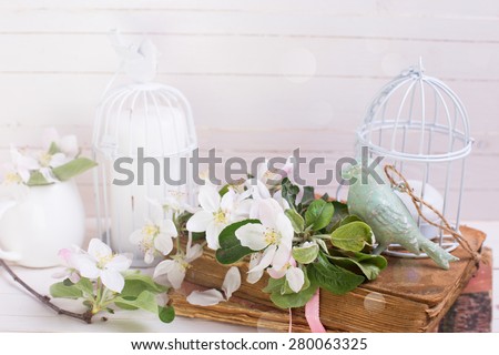 Postcard with apple blossom, decorative bird, old books and candles in decorative bird cages in ray of light on white painted wooden planks. Selective focus.