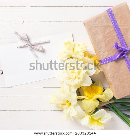 Box with present, spring flowers narcissus  and empty tag for text on white painted wooden planks. Selective focus. Square image.