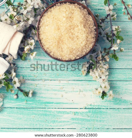 Spa and wellness setting. Sea salt in bowl, towels and  flowering branches of trees in ray of light on turquoise painted wooden background. Selective focus. Square image.