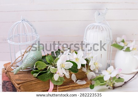 Postcard with apple blossom, decorative bird, old books and candles in decorative bird cages on white painted wooden planks. Selective focus.