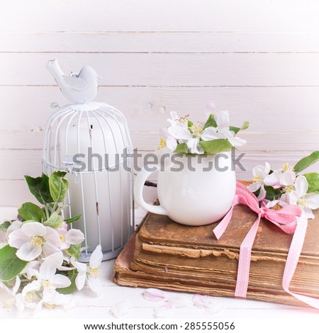 Postcard with apple blossom, old book and candle in decorative bird cages on white painted wooden planks. Selective focus. Square image.