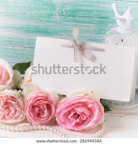 Postcard with sweet roses flowers and empty tag for your text on white painted wooden background against turquoise wall. Selective focus. Square image.