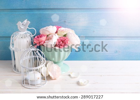 Pastel roses in turquoise vase and candles in ray of light on white wooden  background against blue wall. Place for text. Selective focus.