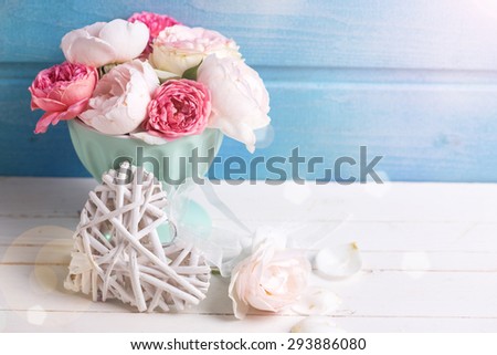 Pastel roses in turquoise vase and decorative heart  in ray of light on white wooden  background against blue wall. Place for text. Selective focus.