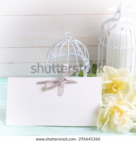 Background with fresh spring flowers, tag and candles in decorative bird cages on turquoise painted planks against white wall. Selective focus is on tag. Square image.