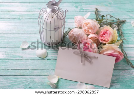Background with fresh roses flowers, candle in decorative bird cage and empty tag on turquoise painted wooden background. Selective focus in on tag.