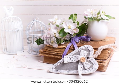 Postcard with apple blossom, decorative heart, old books and candle in decorative bird cage on white painted wooden planks. Selective focus is on apple blossom. Toned image.