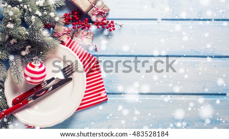 Christmas table setting. White plate, knife and fork, napkin and christmas decorations in white and red colors on blue wooden table. Top view. Drawn snow. Selective focus. Place for text.