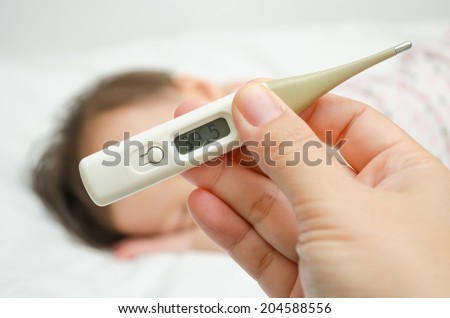 Mother holding thermometer foreground and sick little girl in be