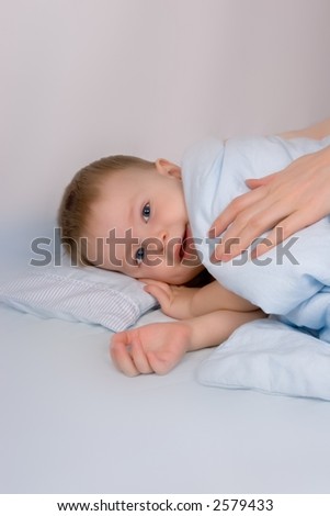 Small boy two years old is going to bed. His mother covers him with a blanket.