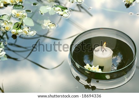 white candle in glass floating in water reflecting sky and flowers of jasmin