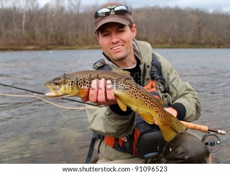 A fly fisherman posing with a Brown Trout with his fly rod and reel, before releasing the fish into the river