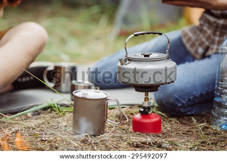 A camping tea kettle on a propane stove next to a metal cup