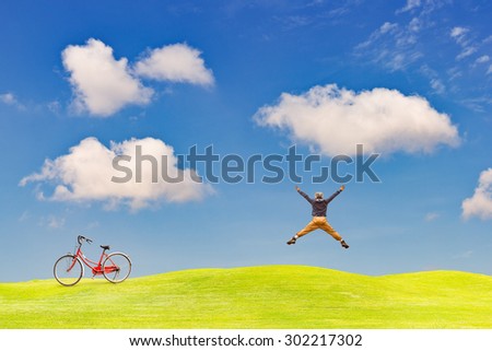 Happy man jumping on green grass field against clear blue sky with an old bicycle on the grass field