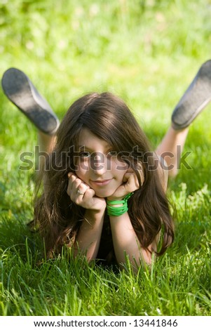 The smiling dark-haired girl-teenager lays on a green lawn in sunlight patches