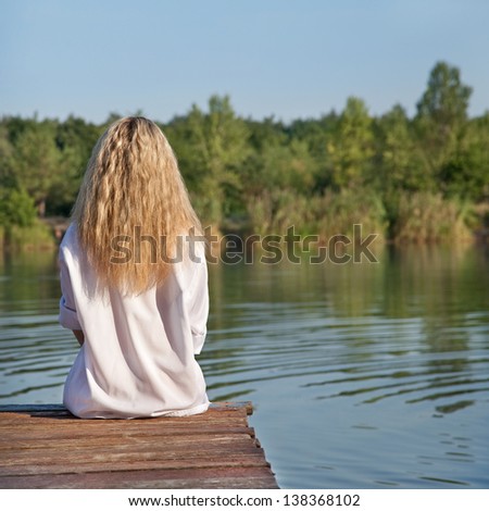 Young girl sitting on pier and looking ahead on river in summer day