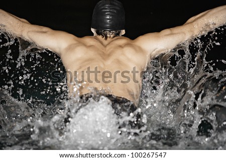 Back view of a male swimmer doing butterfly strokes and splashing water in a swimming pool.
