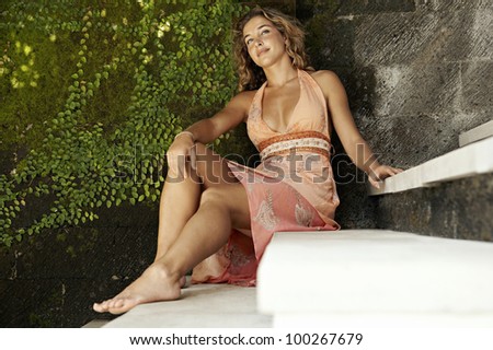 Young woman sitting down on some steps, leaning on a leafy textured wall.
