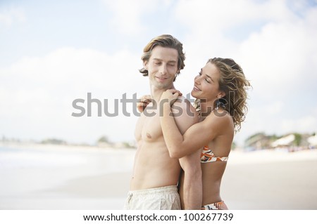 Young attractive woman hugging man from behind while standing on a white sand beach.