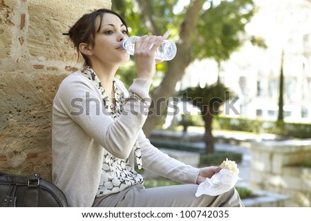 Young businesswoman drinking water from a small plastic bottle while having a lunch break in the park.
