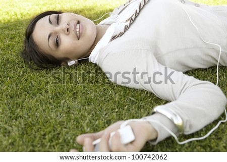 Close up portrait of a  young woman listening to music on her mp4, laying down on green grass in a park.