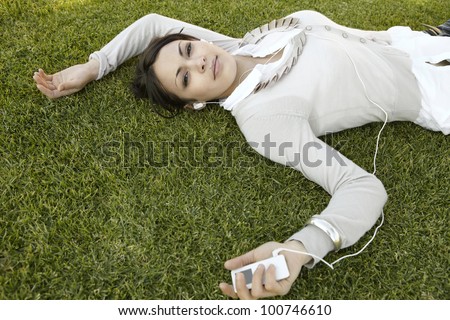 Young woman laying down on green grass, listening to music with headphones.