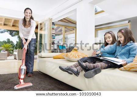 Young mum using a vacuum cleaner while her two twin daughters look at a book in the living room.