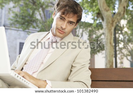 Young businessman using a laptop computer while sitting on a wooden bench in the city.