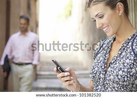 Attractive businesswoman using her cell phone outdoors, while a businessman walks by in the background.