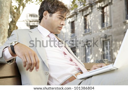 Businessman sitting on a wooden bench using a laptop computer in the city.