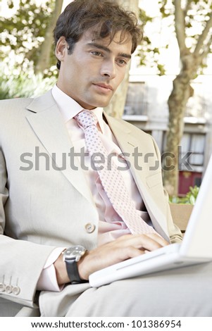 Close up portrait of a businessman sitting on a wooden bench using a laptop computer in the city.