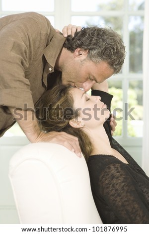 Mature man kissing woman on her forehead while she sits on an armchair in their home's living room.