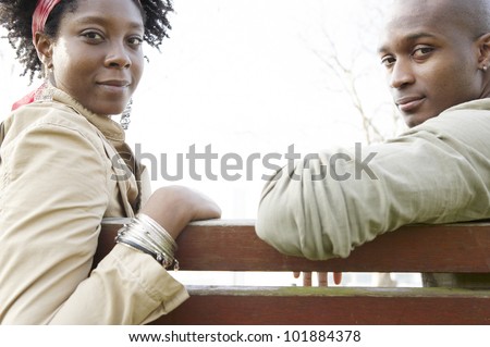 Young couple sitting on a wooden bench, turning around to look at camera with space between them.