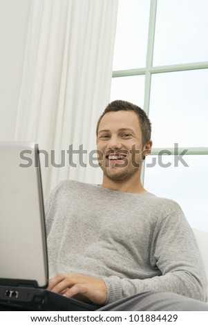 Portrait of a young professional man using a laptop while sitting on a sofa at home, smiling at camera.